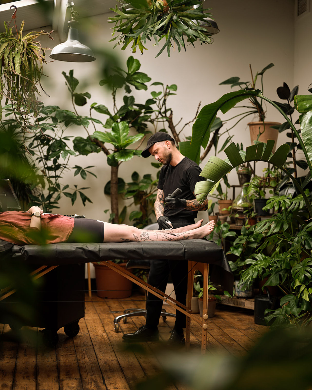 Daniel the Gardener tattooing a reclining client in a plant-filled studio