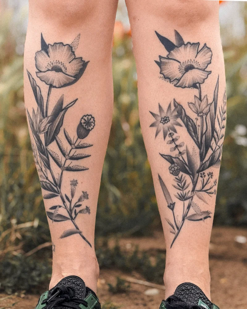 Paired black botanical tattoos on both lower legs, photographed outdoors.