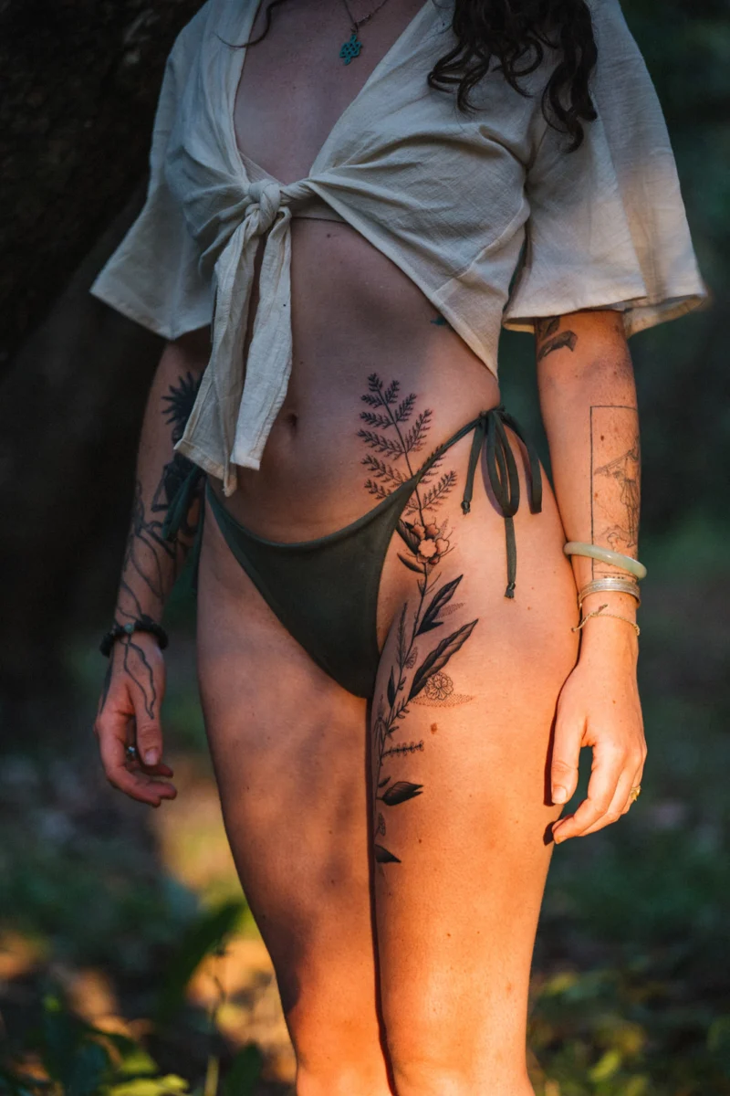 A black botanical tattoo extending from the hip down the thigh, photographed in woodland.