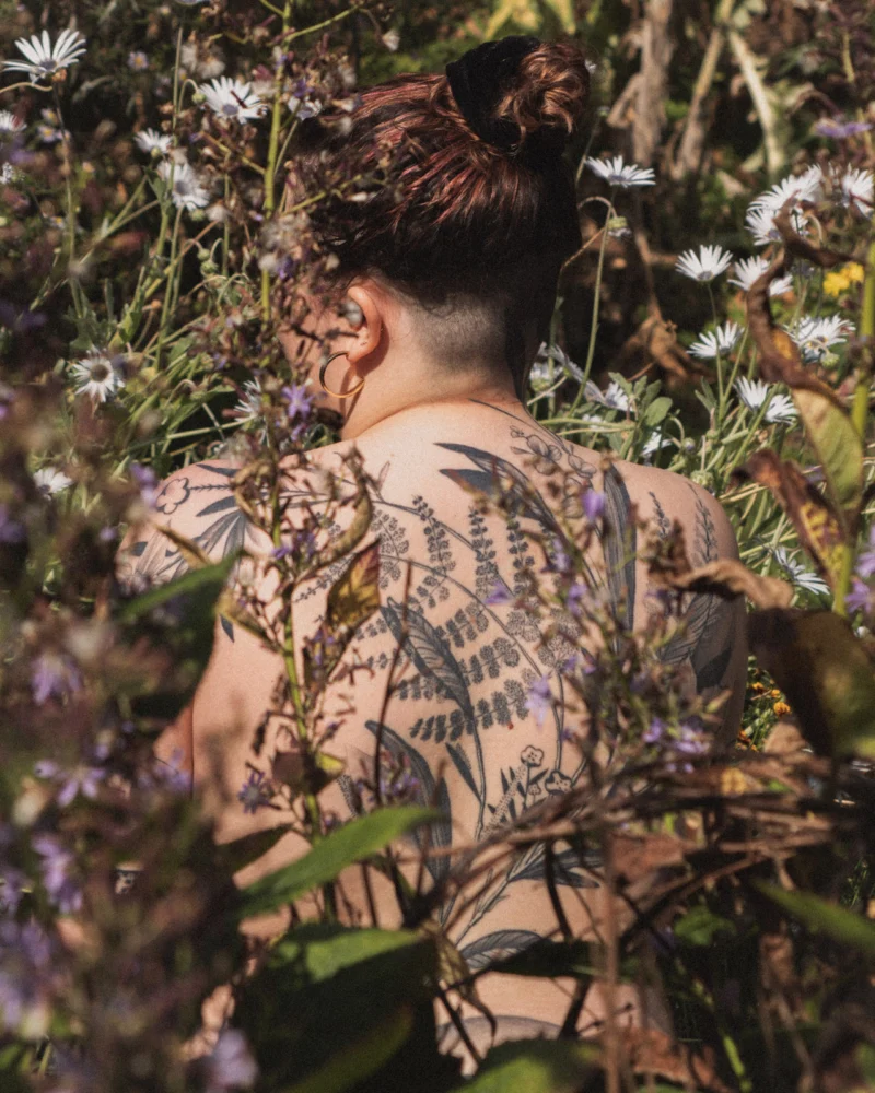 Person with a large botanical back tattoo standing among flowering plants