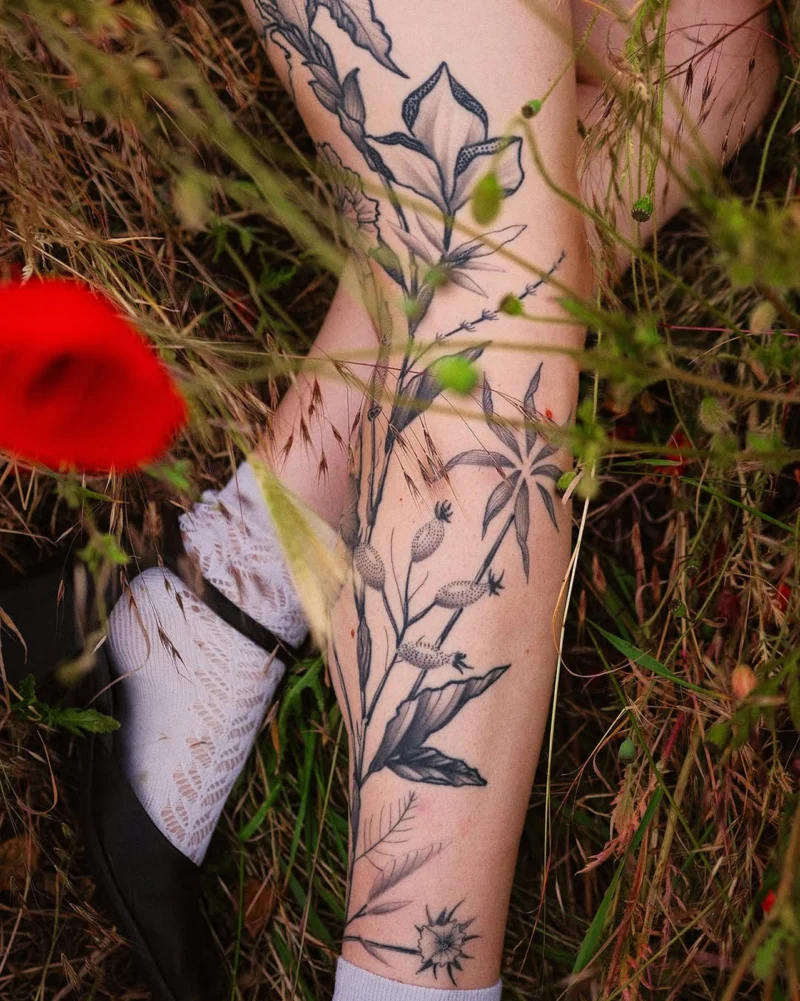 A black botanical leg tattoo photographed among meadow grasses and red poppies.