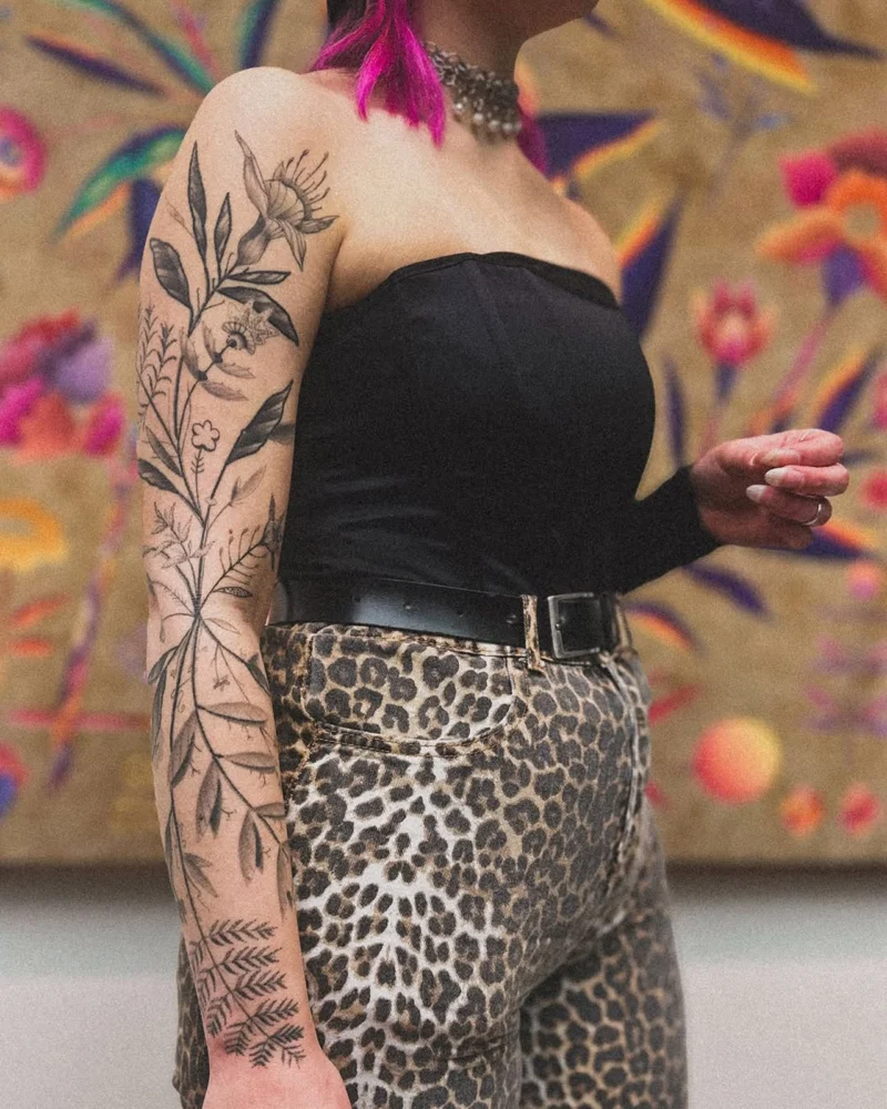 A person with a botanical sleeve tattoo photographed against the FLOWERS exhibition backdrop at Saatchi Gallery.