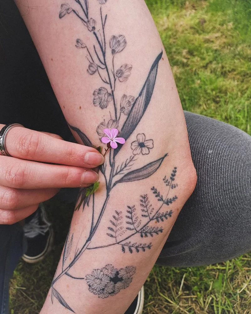 Botanical tattoo on a forearm with a hand holding a small pink flower beside it