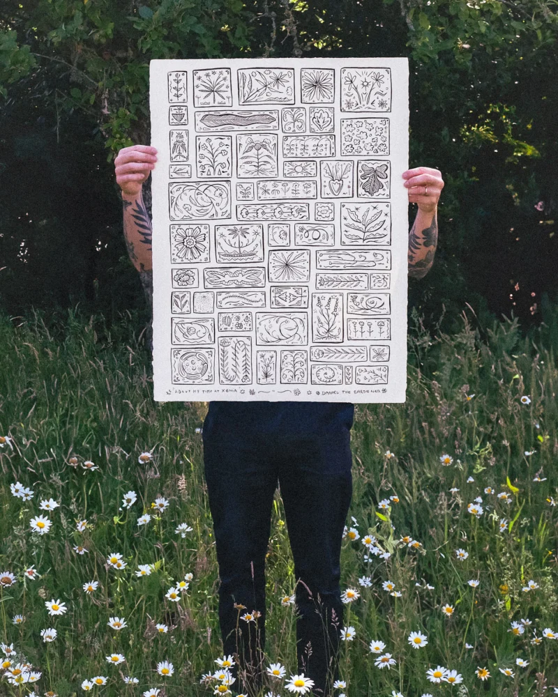 Person holding a large sheet filled with a grid of monochrome botanical drawings in a wildflower meadow
