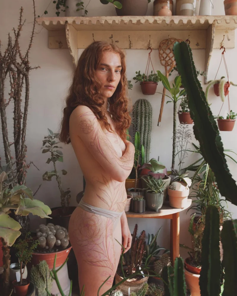 A person with a pink temporary botanical body drawing standing among cacti and houseplants.