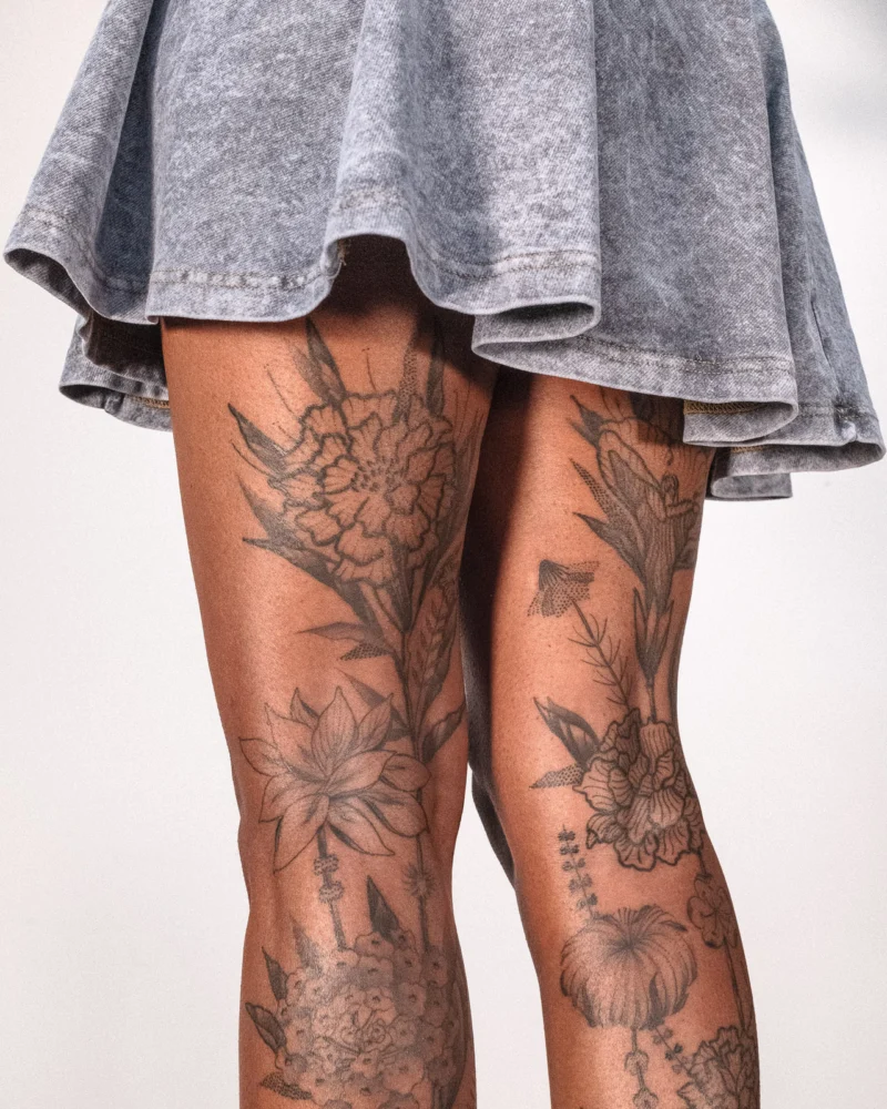 Black botanical tattoos covering both legs, seen from behind beneath a grey skirt