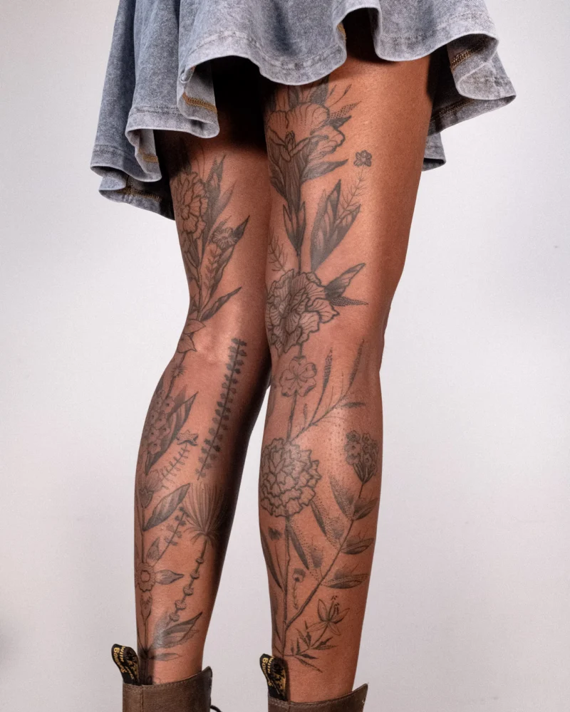 Black botanical tattoos covering both legs, seen from the side beneath a grey skirt